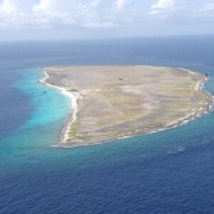Aerial view of Klein Curacao: a flat, sparsely vegetated island with white sand, rocky shores, clear blue waters, small boats, and a central structure under partly cloudy skies. Enjoy it with the Boat Trip Klein Curacao experience.