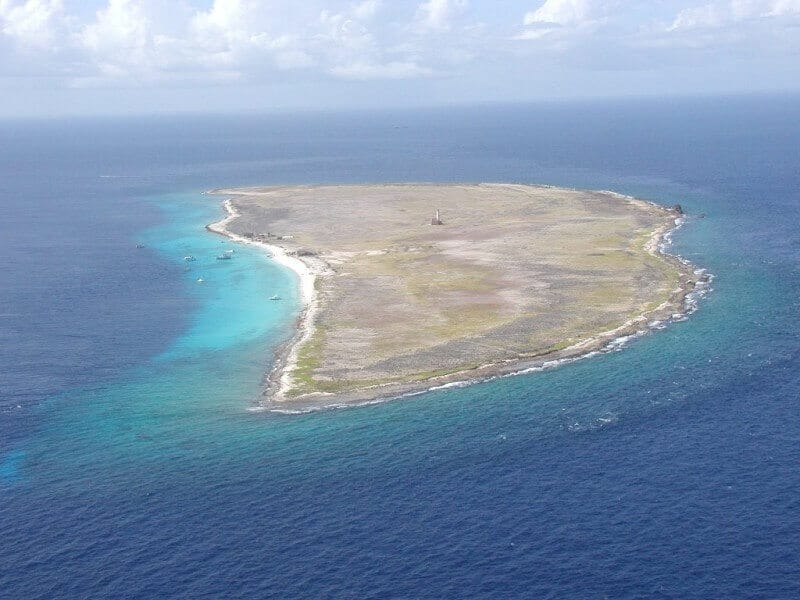 Aerial view of Klein Curacao: a flat, sparsely vegetated island with white sand, rocky shores, clear blue waters, small boats, and a central structure under partly cloudy skies. Enjoy it with the Boat Trip Klein Curacao experience.