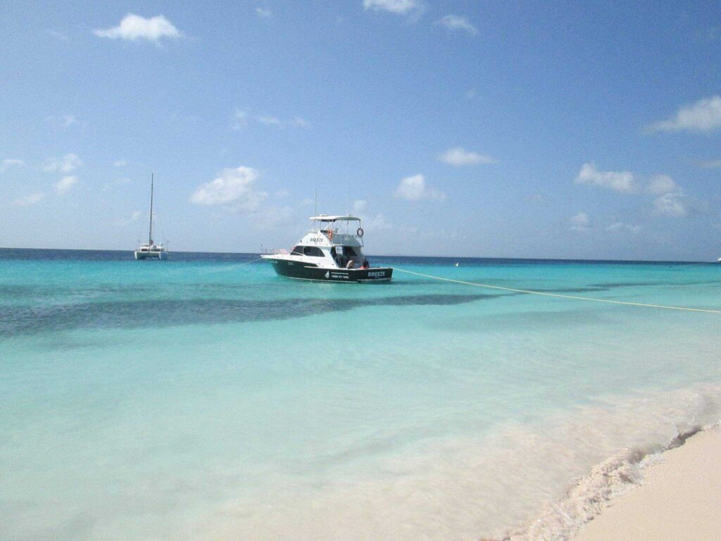 On the Klein Curacao Boat Trip, a small black and white motorboat is anchored near a sandy beach with turquoise water. A sailboat floats in the distance beneath a mostly clear sky, while a rope from the motorboat reaches the shore.