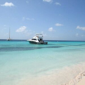 On the Klein Curacao Boat Trip, a small black and white motorboat is anchored near a sandy beach with turquoise water. A sailboat floats in the distance beneath a mostly clear sky, while a rope from the motorboat reaches the shore.