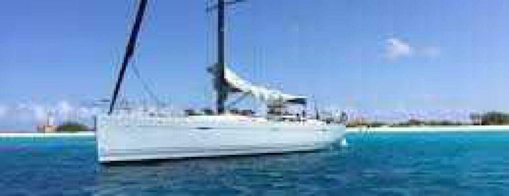 Experience the Klein Curacao Day trip: a white sailboat floats on clear blue water near a sandy shore, under a bright sky with few clouds. The distant coastline and sparse greenery complete the stunning view.