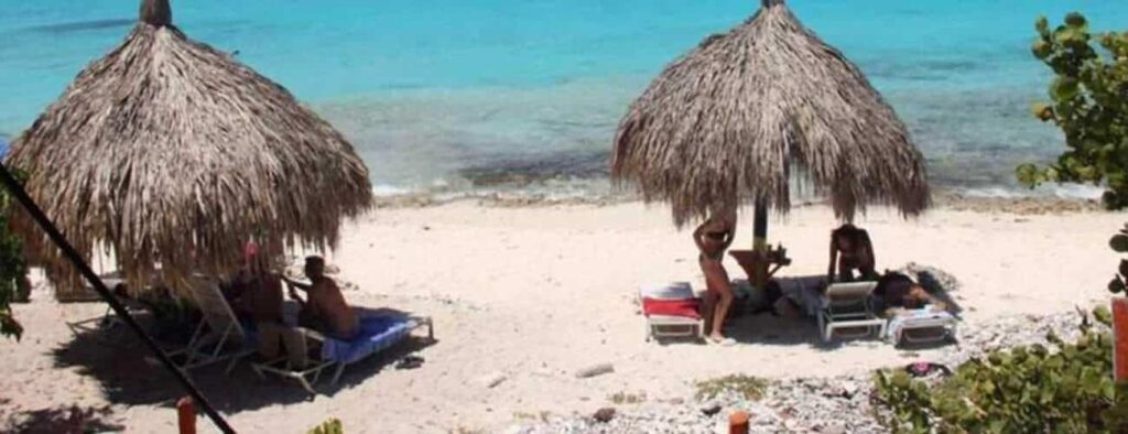 Twee rieten parasols geven schaduw aan mensen die luieren op stoelen op het zandstrand tijdens hun Klein Curacao Reis. De turquoise oceaan is kalm en tropisch groen begrenst dit zonnige, vredige paradijs.