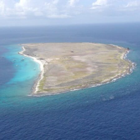 Aerial view from the Klein Curacao trip: a flat, sparsely vegetated island surrounded by clear turquoise waters, deeper blue ocean beyond, small boats offshore, and partly cloudy skies overhead.