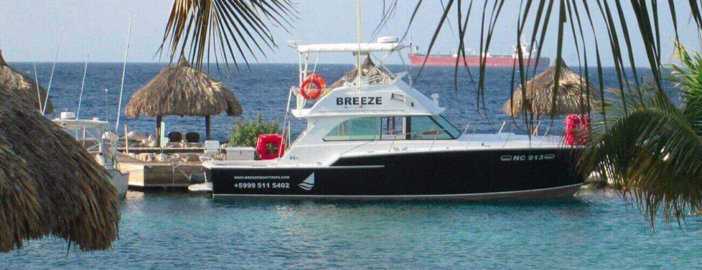 A black and white boat named BREEZE is docked at a tropical marina in Korsou, surrounded by palm trees and thatched-roof huts. The calm blue sea invites you to embark on the Korsou Discovery Boot tour, with a large red ship on the horizon.