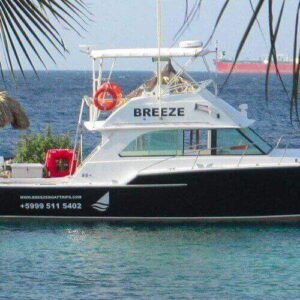 A black and white boat named BREEZE is docked at a tropical marina in Korsou, surrounded by palm trees and thatched-roof huts. The calm blue sea invites you to embark on the Korsou Discovery Boot tour, with a large red ship on the horizon.
