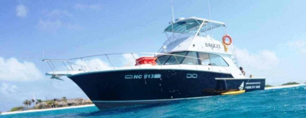 A large blue and white motorboat named BREEZE is anchored near the shore under a sunny sky, ideal for the Korsou Discovery Deluxe Boot tour. Palm-lined tropical land and a bright orange life ring are visible on board in clear blue water.