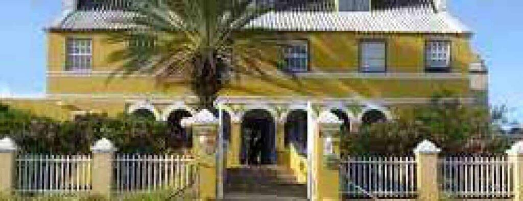 At Landhuis Bloemhof, a yellow colonial-style building with white trim and an arched porch stands behind a white picket fence, beneath a silver sloped roof and bright blue sky, with a tall palm tree and bushes in front.