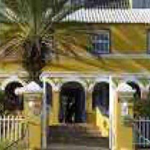 At Landhuis Bloemhof, a yellow colonial-style building with white trim and an arched porch stands behind a white picket fence, beneath a silver sloped roof and bright blue sky, with a tall palm tree and bushes in front.