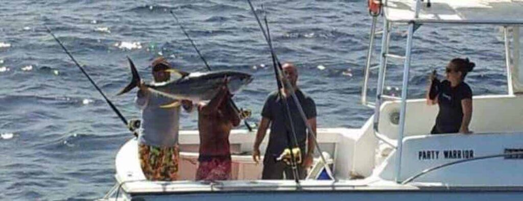 Three people are on a boat named “PARTY WARRIOR” in open water as one lifts a large tuna, showcasing the hands-on "Learn how to fish" experience while others watch and participate.