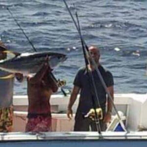 Three people are on a boat named “PARTY WARRIOR” in open water as one lifts a large tuna, showcasing the hands-on "Learn how to fish" experience while others watch and participate.