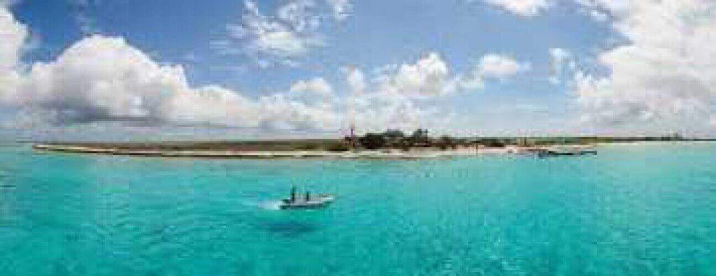 On Little Curaçao, a small sandy island with some trees sits amid clear turquoise water beneath a blue sky with scattered clouds. In the foreground, a boat with two people floats while another boat nears the island’s shoreline.