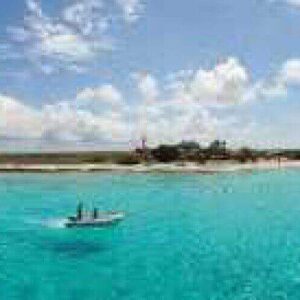 On Little Curaçao, a small sandy island with some trees sits amid clear turquoise water beneath a blue sky with scattered clouds. In the foreground, a boat with two people floats while another boat nears the island’s shoreline.