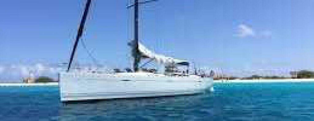 The Local Day trip watersport features a white sailboat with sails down, floating on clear blue water near a sandy coast under a bright sky. Perfect for local outings, guests enjoy sunny weather and watersport activities on deck.