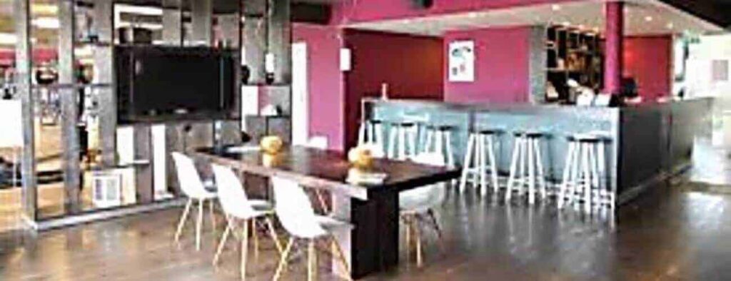 Modern café interior with dark wooden floors, a long table, white chairs, blue bar counter with white stools, and red walls—perfect for gatherings or discussing the Long Term Sport Membership. Shelves and a large TV are on the left wall.