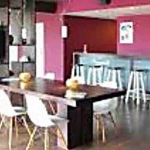 Modern café interior with dark wooden floors, a long table, white chairs, blue bar counter with white stools, and red walls—perfect for gatherings or discussing the Long Term Sport Membership. Shelves and a large TV are on the left wall.