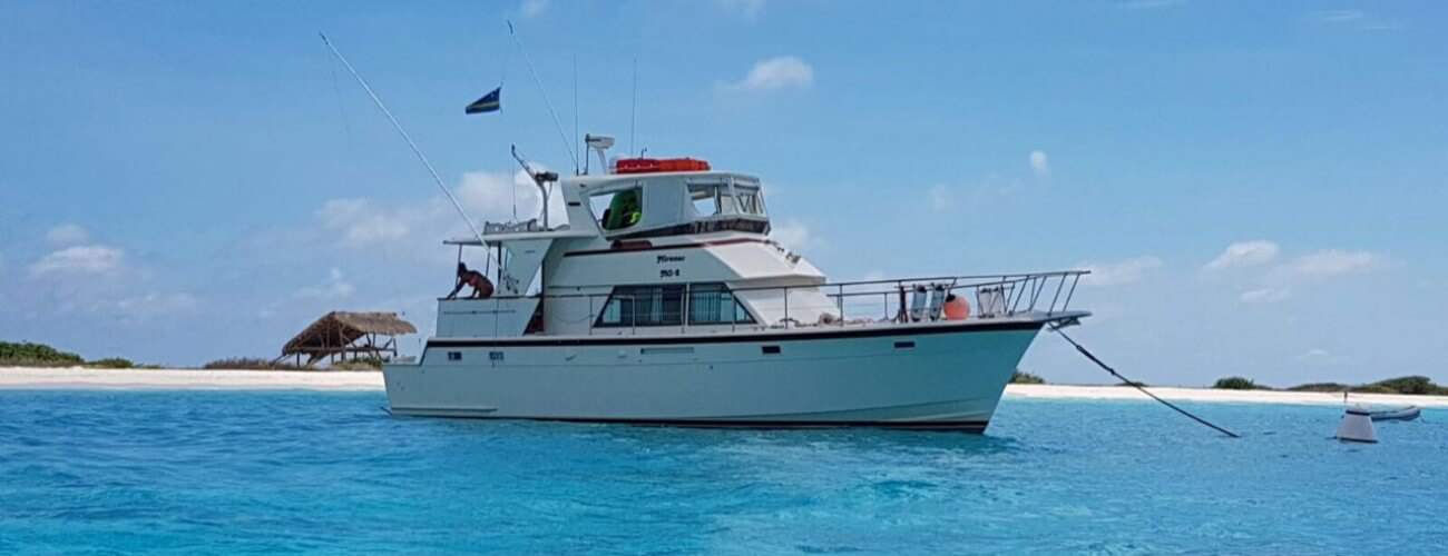 Op Miramar Reis Klein Curaçao ligt een wit motorjacht voor anker in helderblauw water bij een zandeiland met schaars groen en een rieten hut; onder de heldere hemel drijft een boei vlakbij die rust en sereniteit uitstraalt.