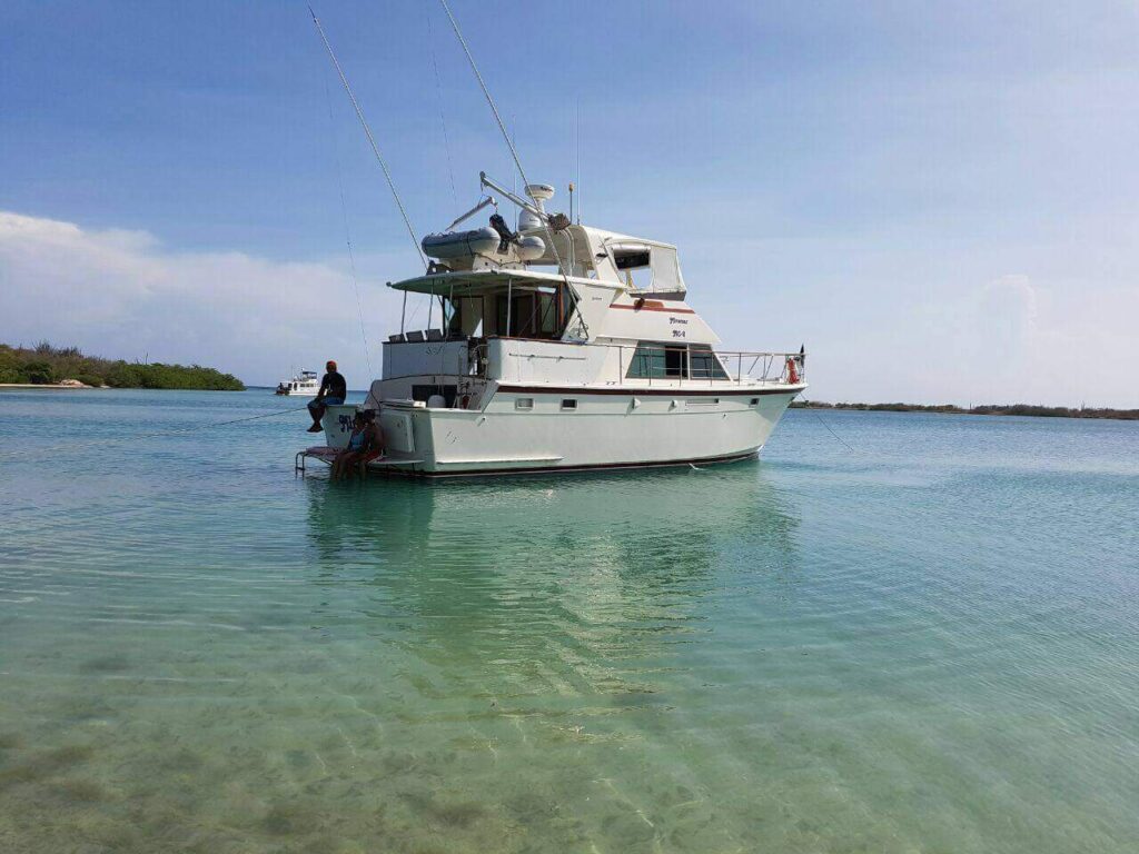 A white motor yacht anchored in clear turquoise water near a small island, with fishing rods on deck and a person enjoying the view, evokes the serene vibe of the Miramar Westpoint Blue Room beneath a vast sky with wispy clouds.