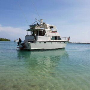 A white motor yacht anchored in clear turquoise water near a small island, with fishing rods on deck and a person enjoying the view, evokes the serene vibe of the Miramar Westpoint Blue Room beneath a vast sky with wispy clouds.