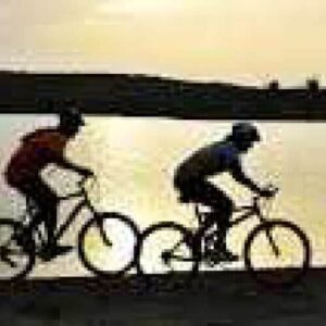 Experience mountain bike tours along a dirt path beside a calm lake at sunset, where two riders’ silhouettes stand out against glowing waters with rolling hills and a clear sky in the background.