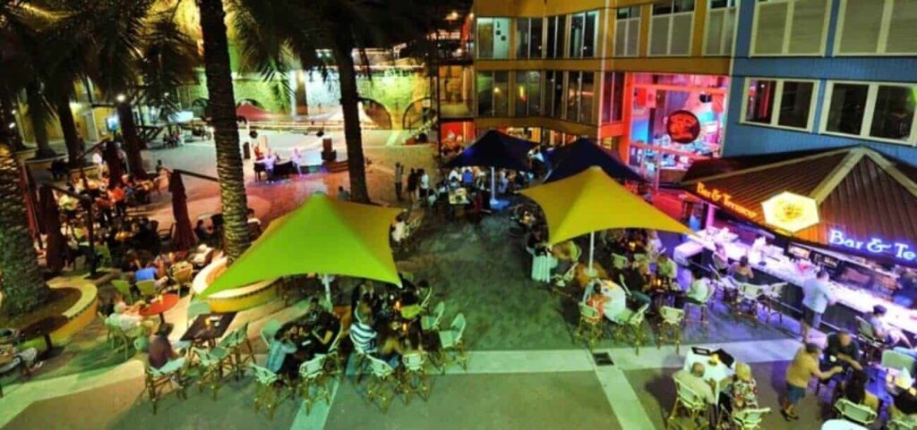 Experience the vibrant Fiesta Night Tour: dine beneath bright yellow and blue canopy tents in an outdoor plaza, surrounded by palm trees, string lights, and a neon-lit bar for a lively, festive atmosphere at night.