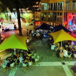 Experience the vibrant Fiesta Night Tour: dine beneath bright yellow and blue canopy tents in an outdoor plaza, surrounded by palm trees, string lights, and a neon-lit bar for a lively, festive atmosphere at night.