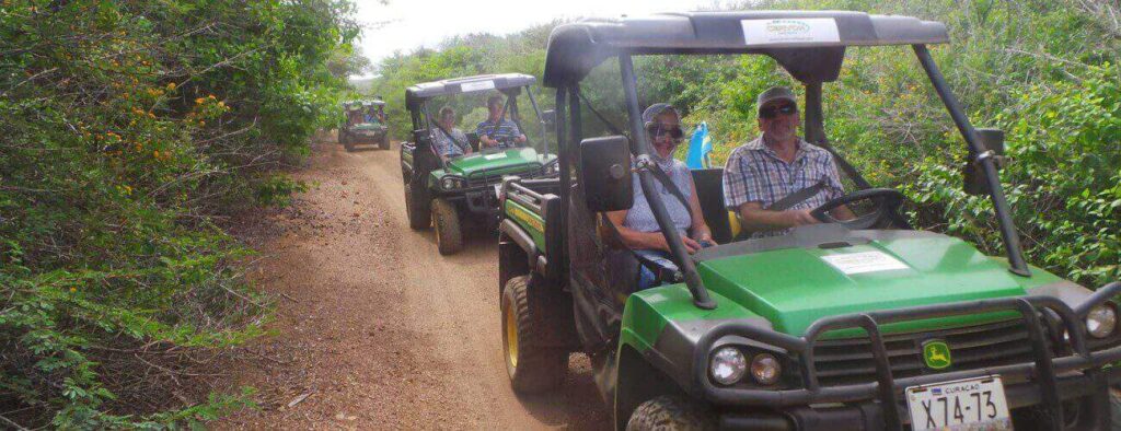 A group explores nature on the Off Road Early Bird Tour, driving green utility vehicles along a narrow dirt trail bordered by dense trees and bushes under partly cloudy skies. They wear sunglasses and casual clothes for this exciting outdoor adventure.