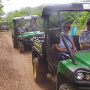 A group explores nature on the Off Road Early Bird Tour, driving green utility vehicles along a narrow dirt trail bordered by dense trees and bushes under partly cloudy skies. They wear sunglasses and casual clothes for this exciting outdoor adventure.