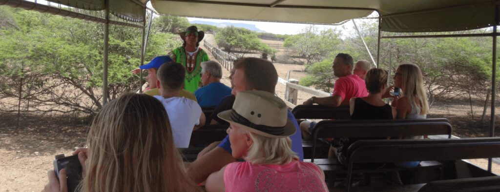 Tourists enjoy a Safari Tour at Ostrich farm in an open-air vehicle with a guide, traveling through dry, bushy terrain under a canopy roof. Many passengers wear hats and summer clothes as they explore the landscape.