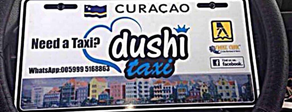 A Curacao taxi sign for "Taxi to Otrobanda" features "Need a Taxi? dushi taxi," a WhatsApp phone number, the Curacao flag, Dutch colonial building images, and local taxi logos on the right.