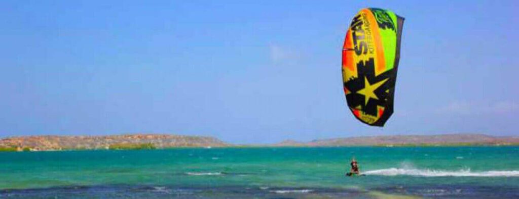 Enjoy Package Private Lessons - Kitesurfing as you glide on turquoise water under a clear blue sky, guided by experts along scenic low hills and a rocky shore in the distance.