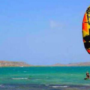 Enjoy Package Private Lessons - Kitesurfing as you glide on turquoise water under a clear blue sky, guided by experts along scenic low hills and a rocky shore in the distance.