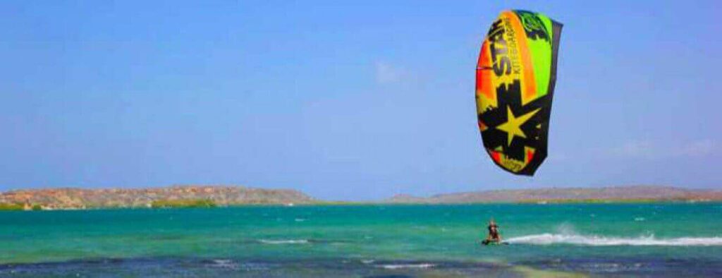 A person takes Package Semi Private Lessons - Kitesurfing, riding on turquoise water under a clear sky as a mostly yellow, orange, and black kite with stars flies high and rocky islands appear on the horizon.