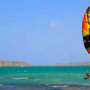 A person takes Package Semi Private Lessons - Kitesurfing, riding on turquoise water under a clear sky as a mostly yellow, orange, and black kite with stars flies high and rocky islands appear on the horizon.