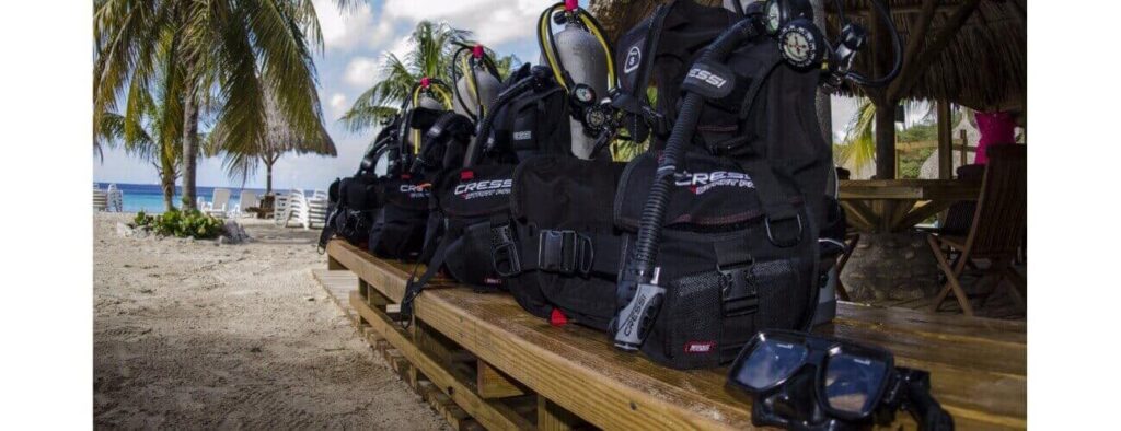 Black buoyancy control devices with regulators and masks are lined up on a bench under a thatched hut, surrounded by palm trees and ocean views, ready for the next PADI Dive Master Course.