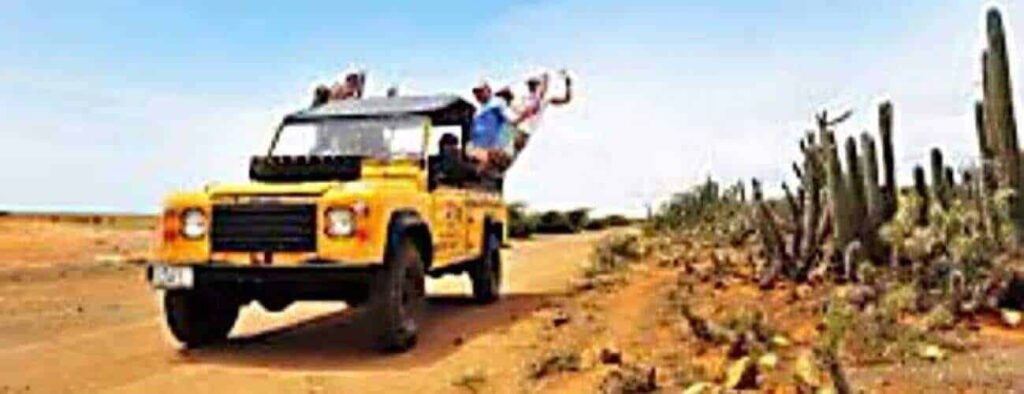 A Pickup safari full vehicle drives down a dusty desert road lined with tall cacti and rocks. Several people smile and wave from the open roof under a clear blue sky.