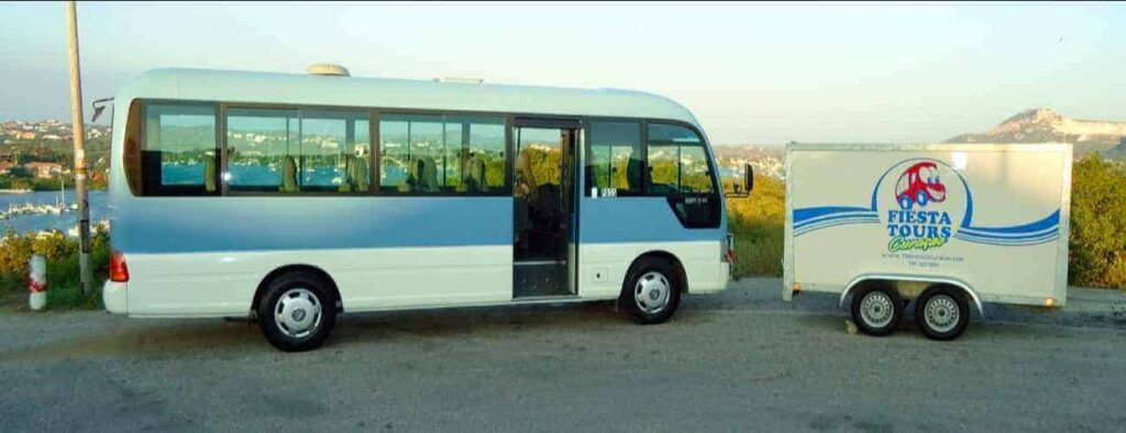 A small blue and white bus for Private Customized Tours is parked roadside with a matching enclosed trailer labeled Fiesta Tours. The open door welcomes you, set against a scenic backdrop of greenery, hills, and water.