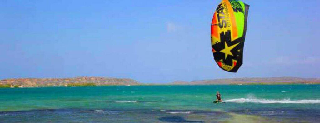 Enjoy a Private Lesson - Kitesurfing as you glide on turquoise water under a clear sky. A vibrant yellow, green, and black kite soars above while waves ripple behind you, with rocky low islands in the background.