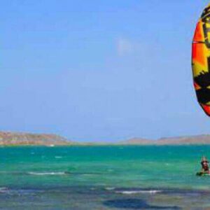 Enjoy a Private Lesson - Kitesurfing as you glide on turquoise water under a clear sky. A vibrant yellow, green, and black kite soars above while waves ripple behind you, with rocky low islands in the background.