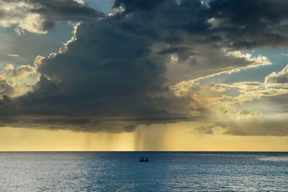A vast ocean under a dramatic Curacao sky with large, dark storm clouds releasing rain in the distance. The sun sets behind the clouds, casting golden light. A small boat is silhouetted against the horizon in the calm water.
