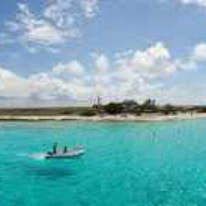 Two people in a small boat float on clear turquoise water near a sandy island with sparse plants, while another Rent a Yacht vessel is docked by the shore under a sky filled with fluffy clouds.