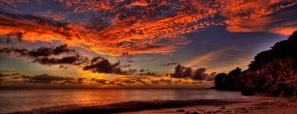 The Romantic Sunset Sail captures a vivid sunset over calm ocean waters with dramatic orange-red clouds, rocky cliffs and trees silhouetted at the edge, and a sailboat drifting by the glowing sun reflected on water and sand.