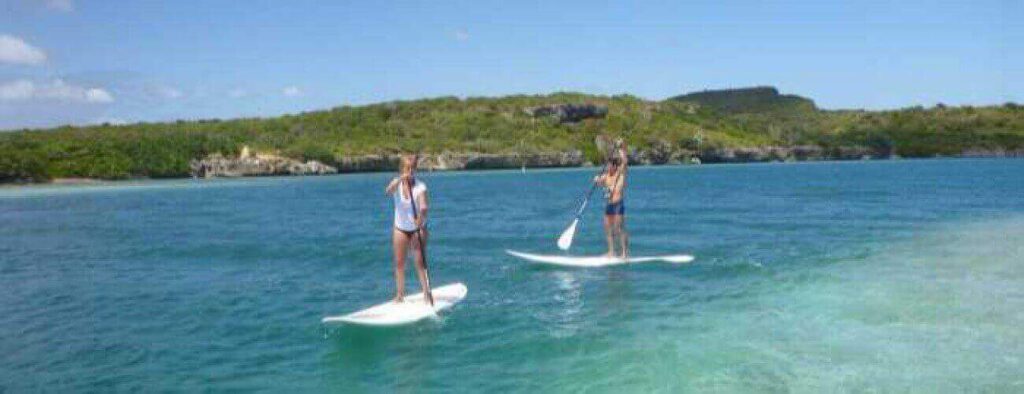 Two people paddle on clear blue water during the Round the Caracasbay Peninsula Tour, with lush green hills in the background—a scenic and tranquil adventure under a bright, partly cloudy sky.