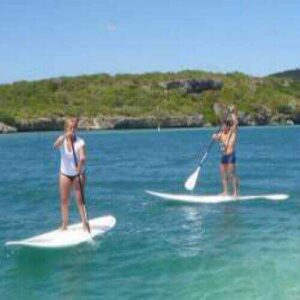 Two people paddle on clear blue water during the Round the Caracasbay Peninsula Tour, with lush green hills in the background—a scenic and tranquil adventure under a bright, partly cloudy sky.