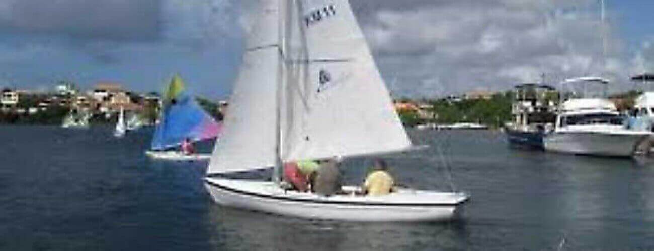 A Sailing course one person dinghy glides with two sailors across calm water. Colorful dinghies and yachts are in the background, with houses and greenery lining the shore under a partly cloudy sky.