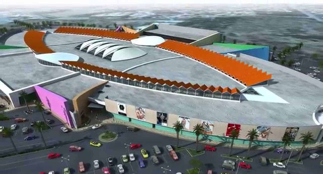 Aerial view of a large, modern Curacao shopping mall with a unique, oval-shaped design, colorful exterior walls, and orange zigzag roof accents. The parking lot below is filled with cars and bordered by palm trees; the surrounding area is blurred.
