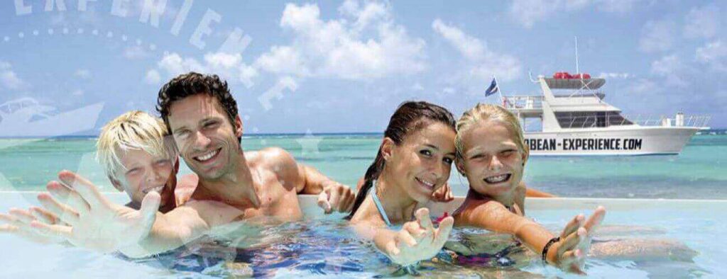 A smiling family of four—two adults and two kids—enjoys a clear, shallow pool, reaching toward the camera. Behind them, a white boat labeled SEAFISHING floats on turquoise sea under a bright blue sky.