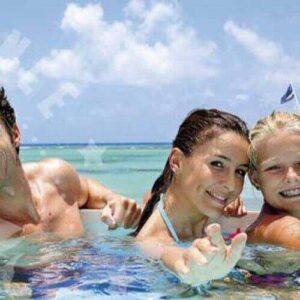 A smiling family of four—two adults and two kids—enjoys a clear, shallow pool, reaching toward the camera. Behind them, a white boat labeled SEAFISHING floats on turquoise sea under a bright blue sky.