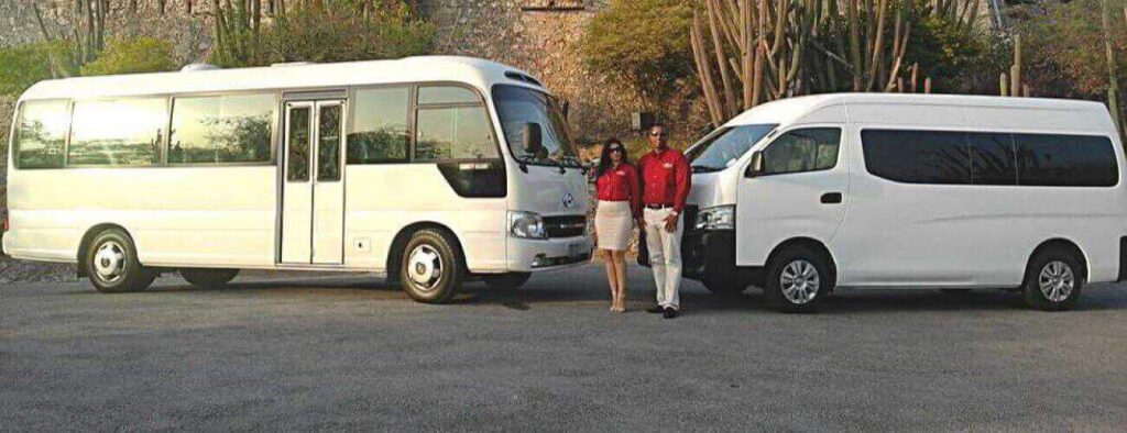 Two white vans for Shared Airport Transfer Zone B are parked outdoors. Two people in red shirts and light-colored bottoms stand smiling in front of the smaller van, with shrubs and trees behind them.