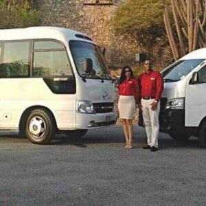 Two white vans for Shared Airport Transfer Zone B are parked outdoors. Two people in red shirts and light-colored bottoms stand smiling in front of the smaller van, with shrubs and trees behind them.