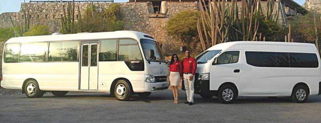 Two people in red shirts and light bottoms stand between a white minibus and van, awaiting their Shared Airport Transfer Zone C amid sunny stone walls, desert plants, and a partially visible building above the wall.